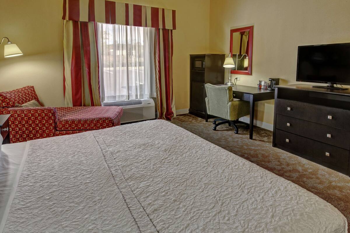 A hotel room with a large bed, a desk with a chair, a television, a dresser, and a window with curtains.