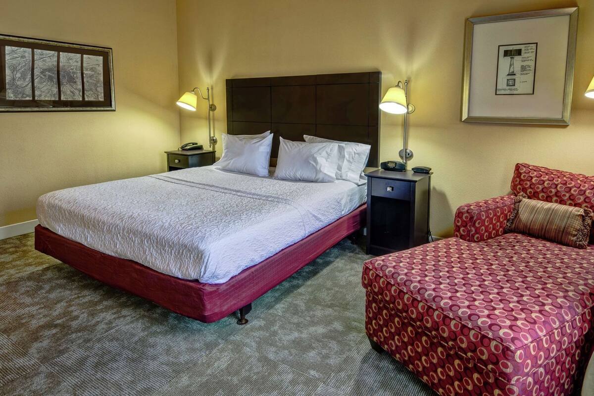 A hotel room with a bed, a bedside table, a lamp, a chair, and a framed picture on the wall.