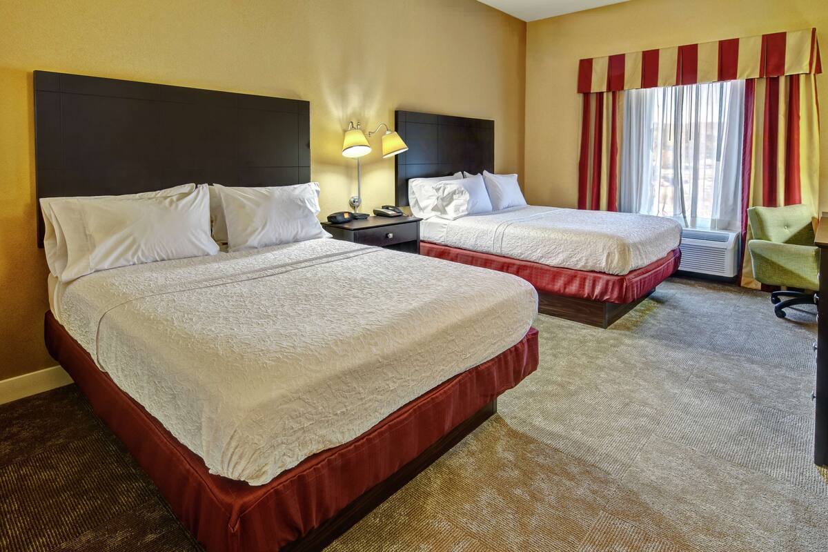 A hotel room with two beds, a nightstand, a lamp, a chair, and a window with curtains.