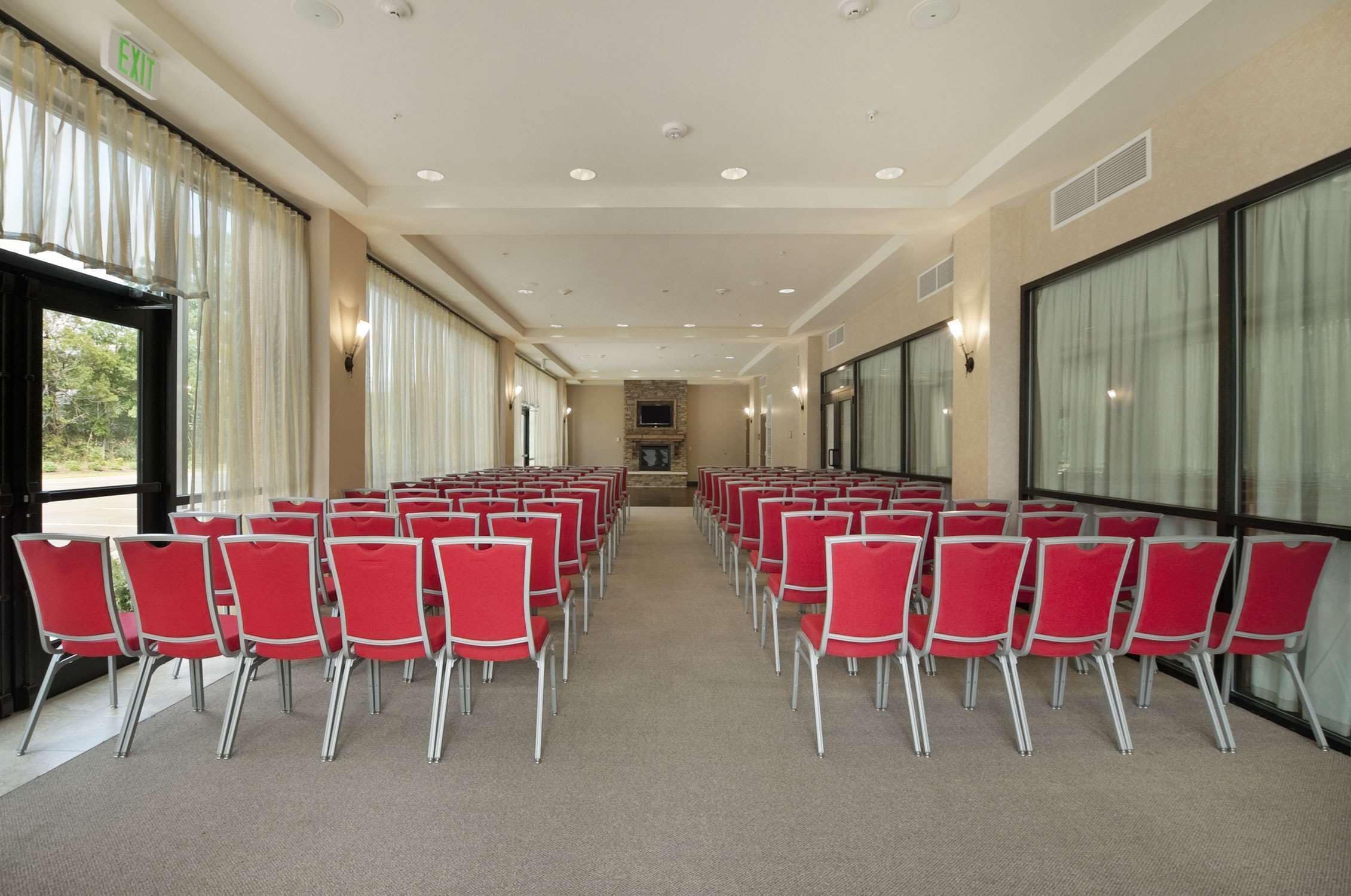 Une salle de conférence avec des rangées de chaises rouges, de grandes fenêtres et un panneau indiquant la sortie.
