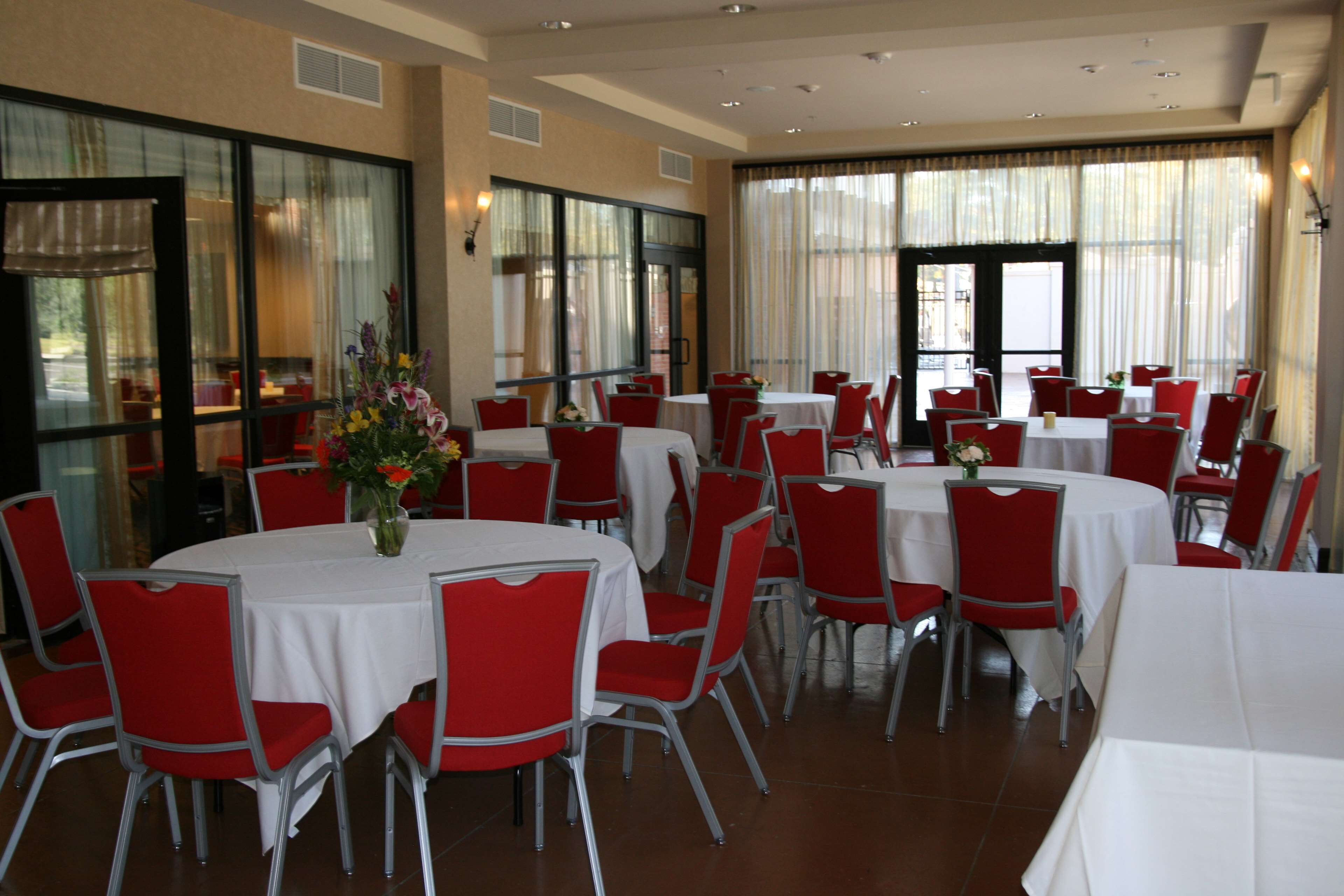 Un espace repas avec des tables rondes et des chaises rouges, de grandes fenêtres avec des rideaux et une vue sur l’extérieur.