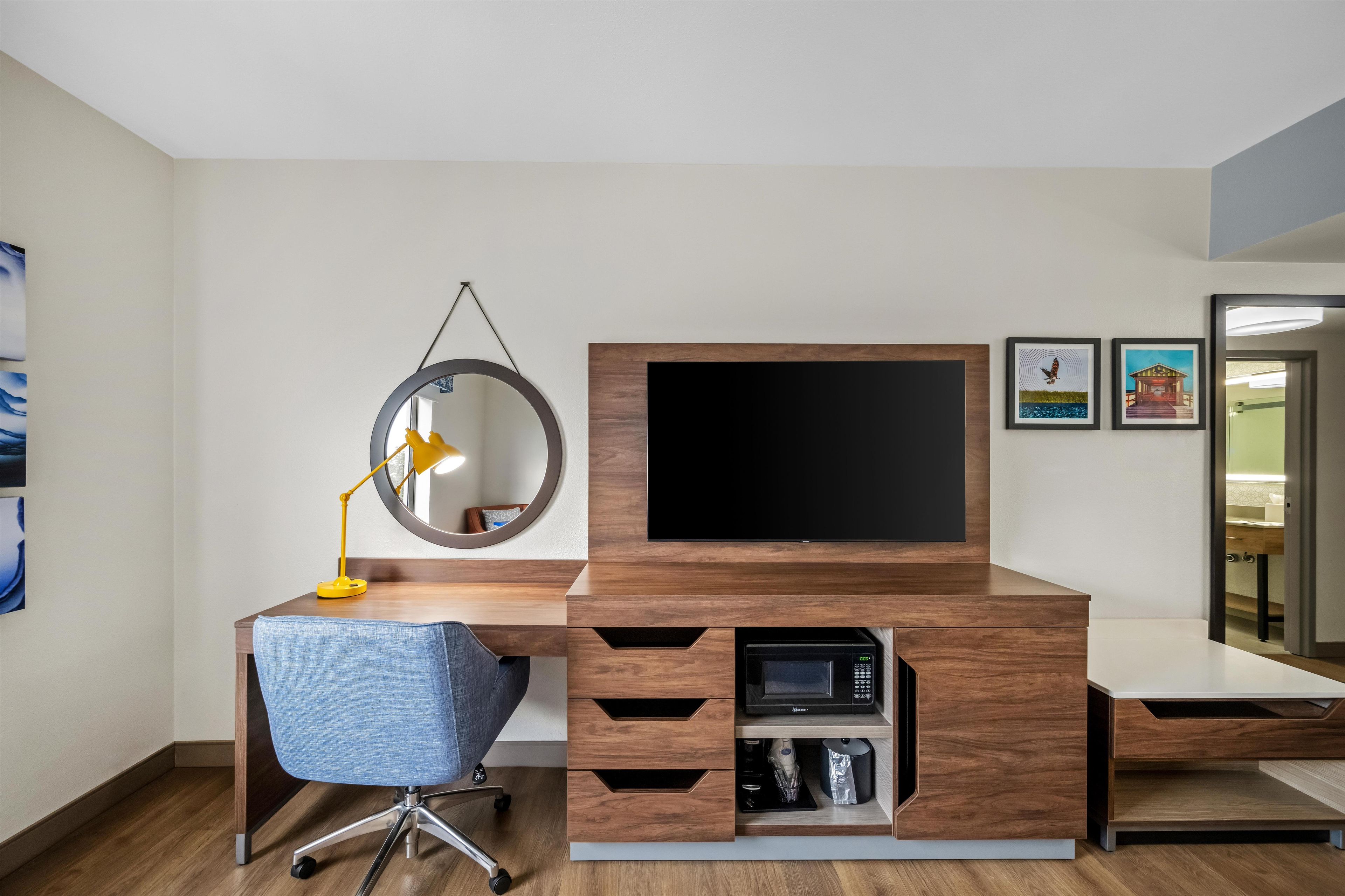 Un espacio de oficina moderno con un escritorio de madera, una silla de oficina azul, un espejo redondo, un televisor plano de pantalla montado y un microondas.
