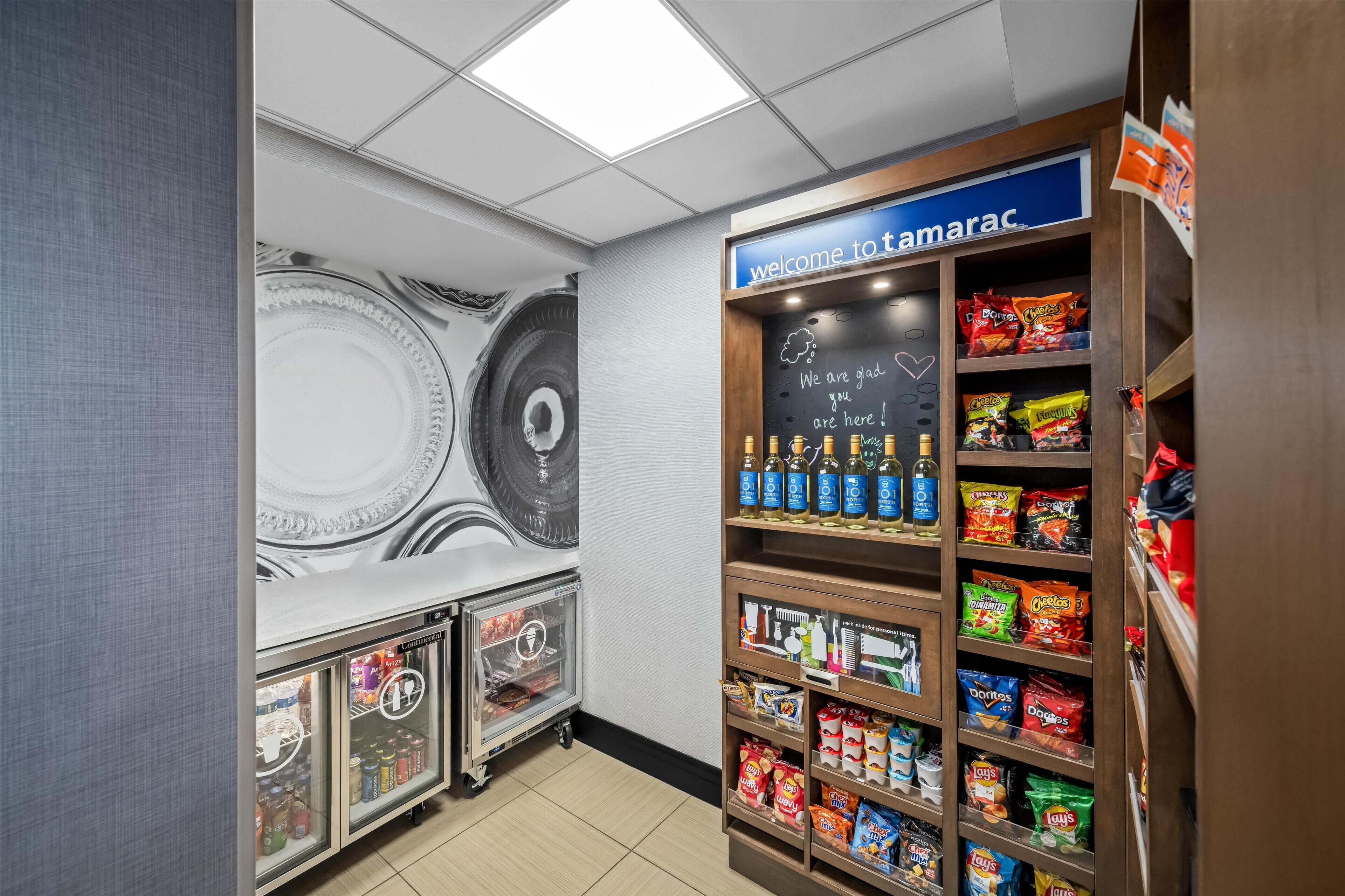 A snack area with a 'welcome to tamarac' sign, refrigerated drinks, and shelves stocked with chips and snacks.