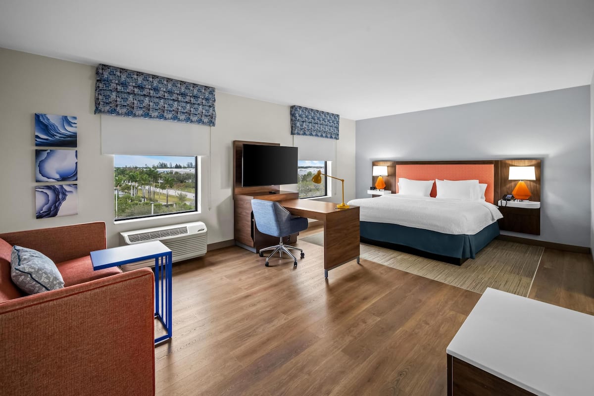Habitación de hotel con cama, escritorio, silla, televisor y ventanas amplias.