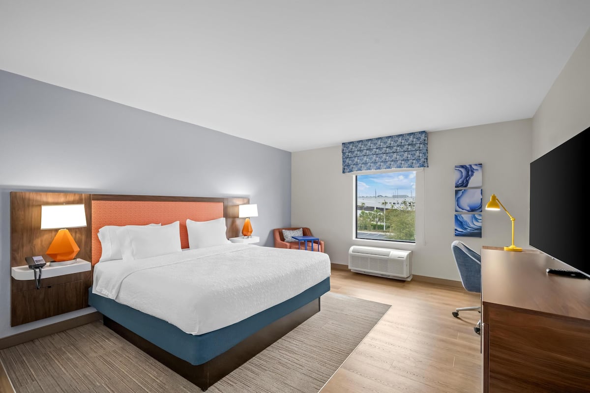 Habitación de hotel con cama, mesitas de noche, escritorio, silla, televisor y ventana con vistas, además de obras de arte en la pared.