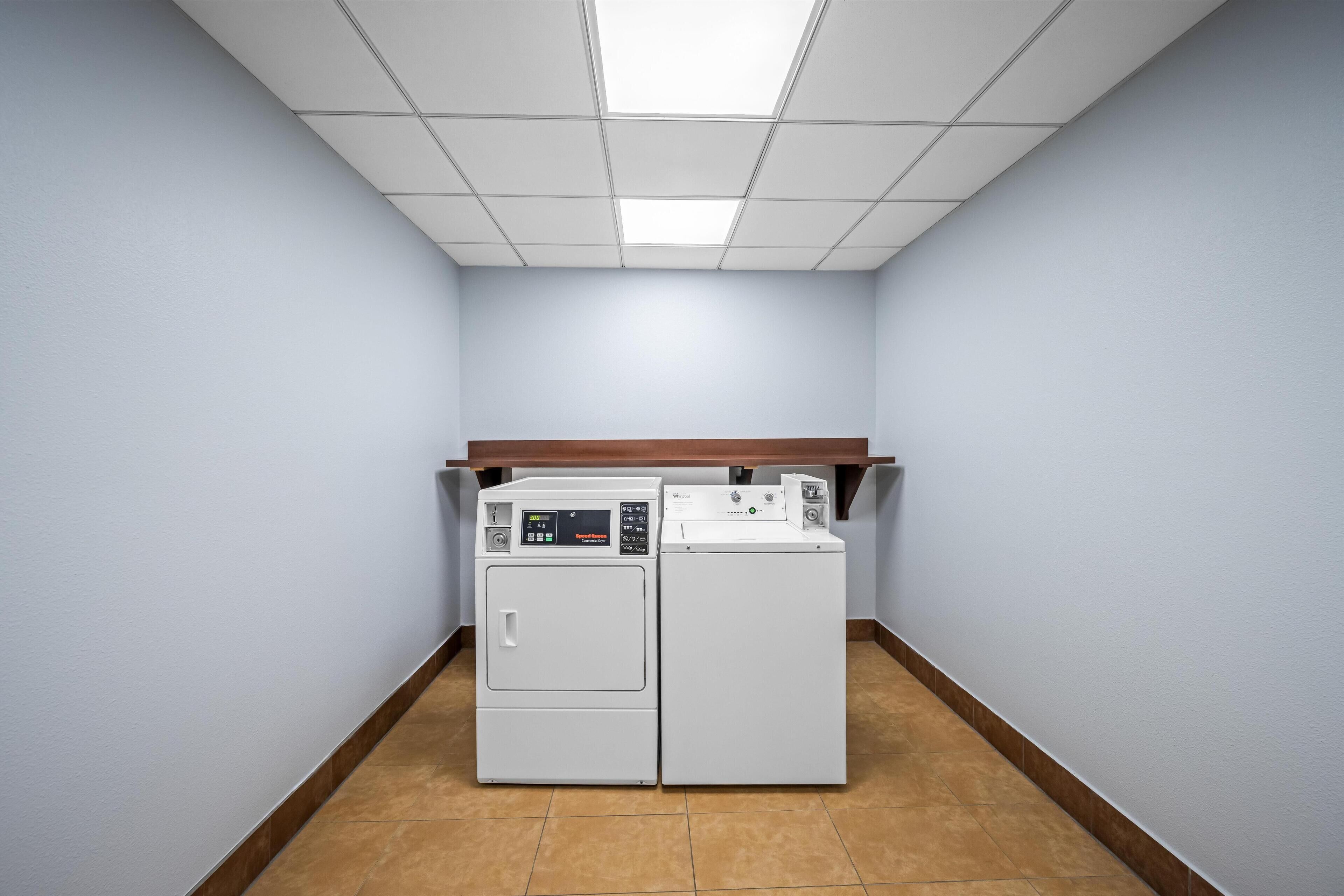 A laundry room with two stacked washers and dryers, a wooden shelf above, and a tiled floor.
