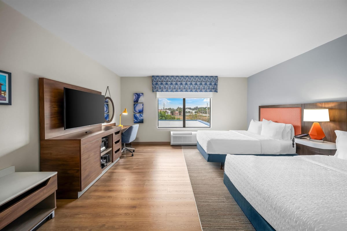 Habitación de hotel con un televisor grande, un escritorio, una silla, una cama, una mesita de noche y un ventanal con vistas al exterior.