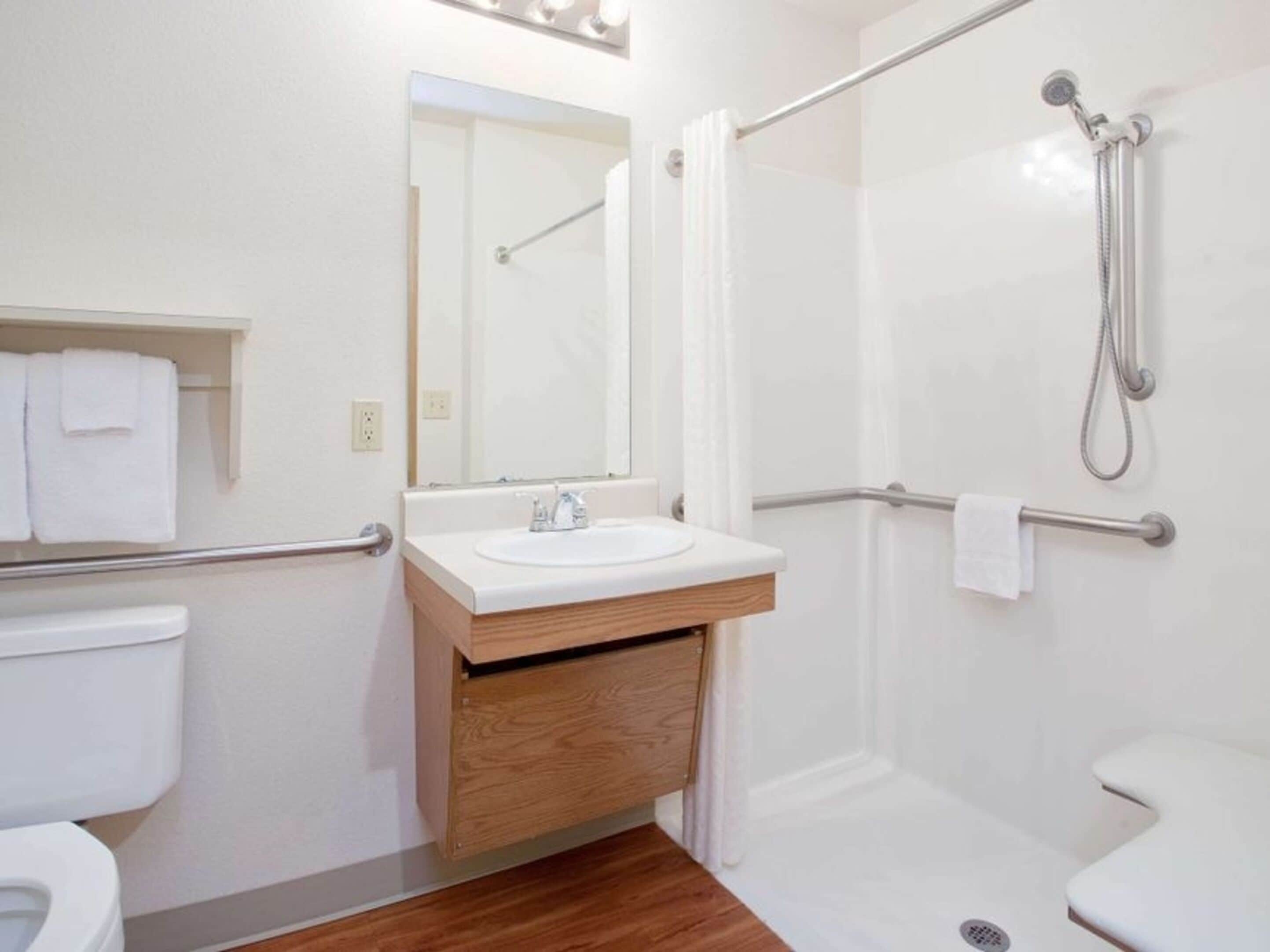 Un baño moderno con lavamanos, espejo y retrete blancos. Incluye una ducha con cabezal de ducha manual y barra de apoyo.