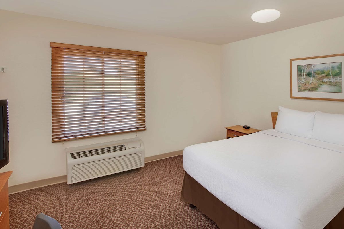 A hotel room with a large bed, a window with blinds, a heater, a nightstand, and a painting on the wall.