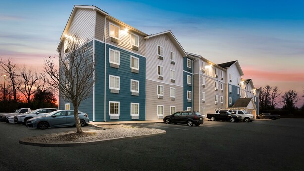A multi-story hotel with a parking lot, featuring a mix of light and dark exterior colors, and a clear sky in the background. - WoodSpring Suites Lexington (Lexington)