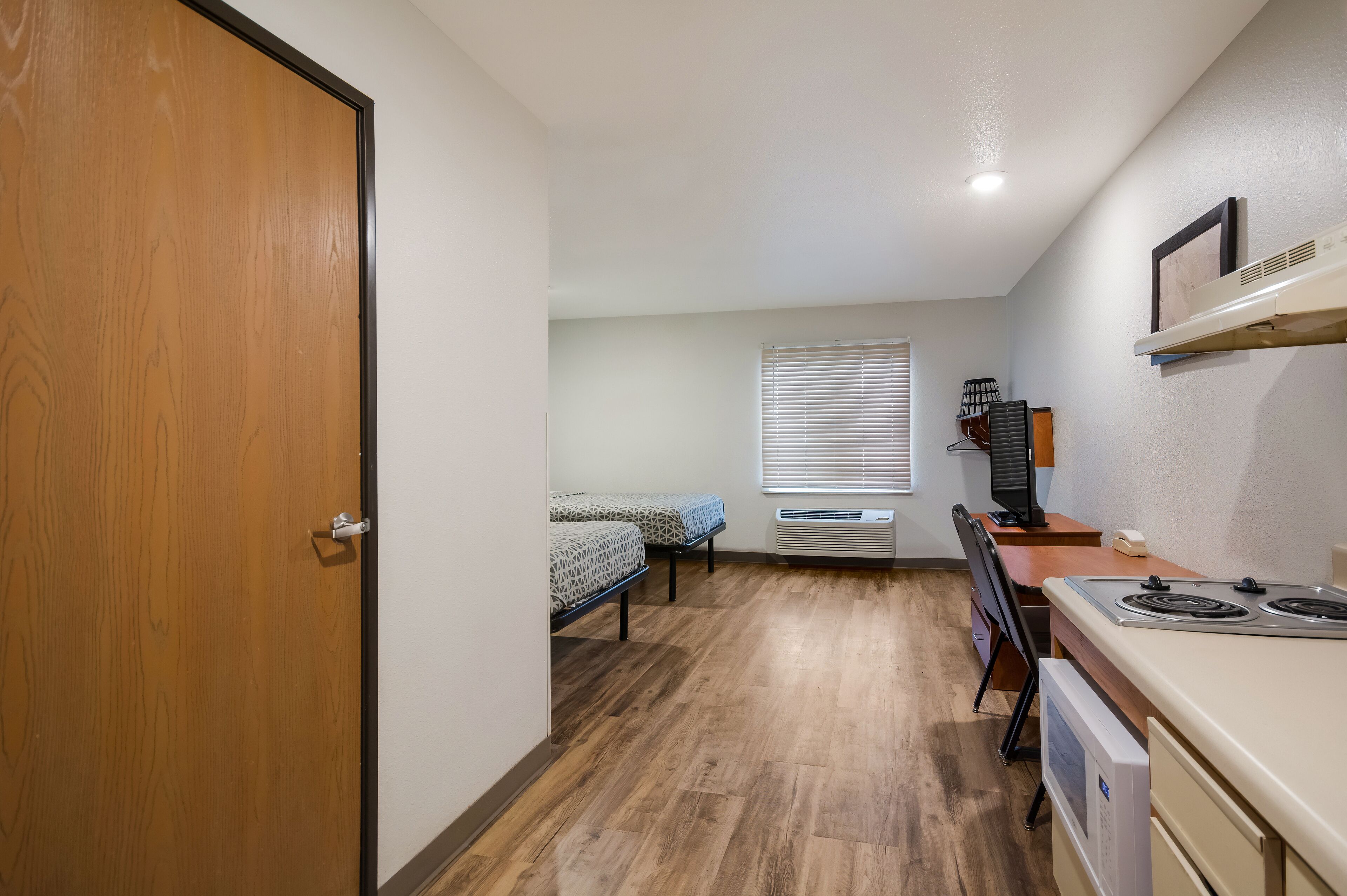 Quarto compacto com porta de madeira, cama com roupa de cama estampada, escrivaninha com computador e cozinha compacta com fogão.