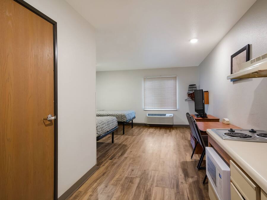 Quarto compacto com porta de madeira, cama com roupa de cama estampada, escrivaninha com computador e cozinha compacta com fogão.