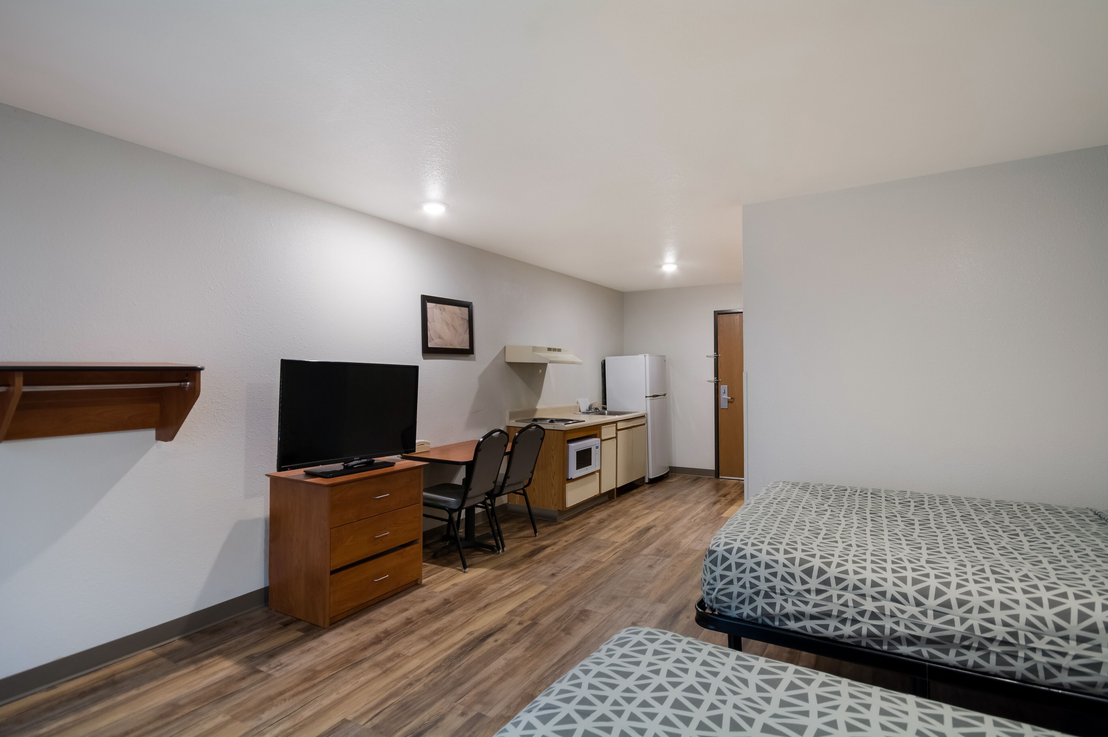 A compact bedroom with a bed, a desk, a chair, a television, and a small kitchenette.