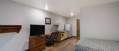 A compact bedroom with a bed, a desk, a chair, a television, and a small kitchenette.