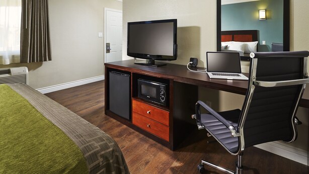 A hotel room with a bed, a desk with a laptop, a television, a microwave, and a chair. - American Inn (Downey)