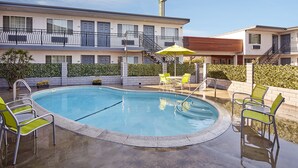 A swimming pool with a sunshade, lounge chairs, and a building with a sign that reads "Stuart Inn." - Studio Inn and Suites at Promenade Downey (Downey)