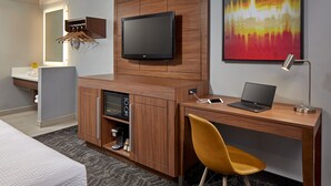 A modern hotel room with a wooden desk, a flat-screen TV, a laptop, a yellow chair, a coffee machine, and a mirror with a light. - Studio Inn and Suites at Promenade Downey (Downey)