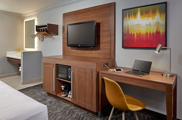 A modern hotel room with a wooden desk, a flat-screen TV, a laptop, a yellow chair, a coffee machine, and a mirror with a light. - Studio Inn and Suites at Promenade Downey (Downey)