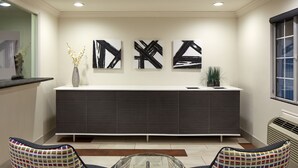 A reception area with a modern reception desk, two patterned chairs, a vase with flowers, and three abstract wall art pieces. - Studio Inn and Suites at Promenade Downey (Downey)
