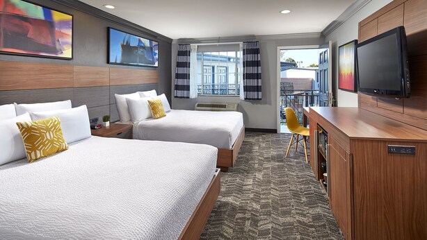 A hotel room with two beds, a desk, a TV, and a view of the outdoors. - Studio Inn and Suites at Promenade Downey (Downey)