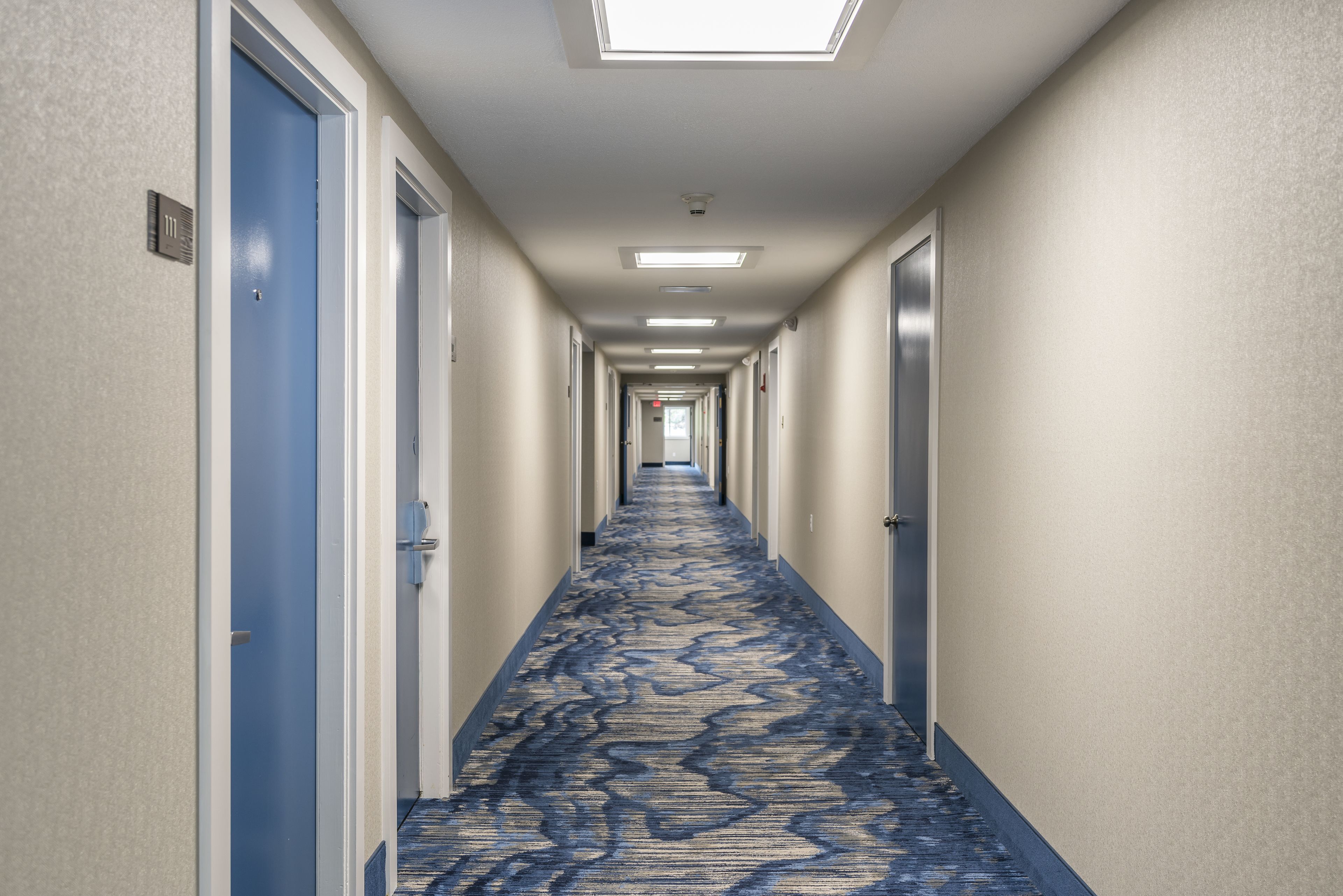 A hotel corridor with blue carpet, doors on both sides, and overhead lighting.