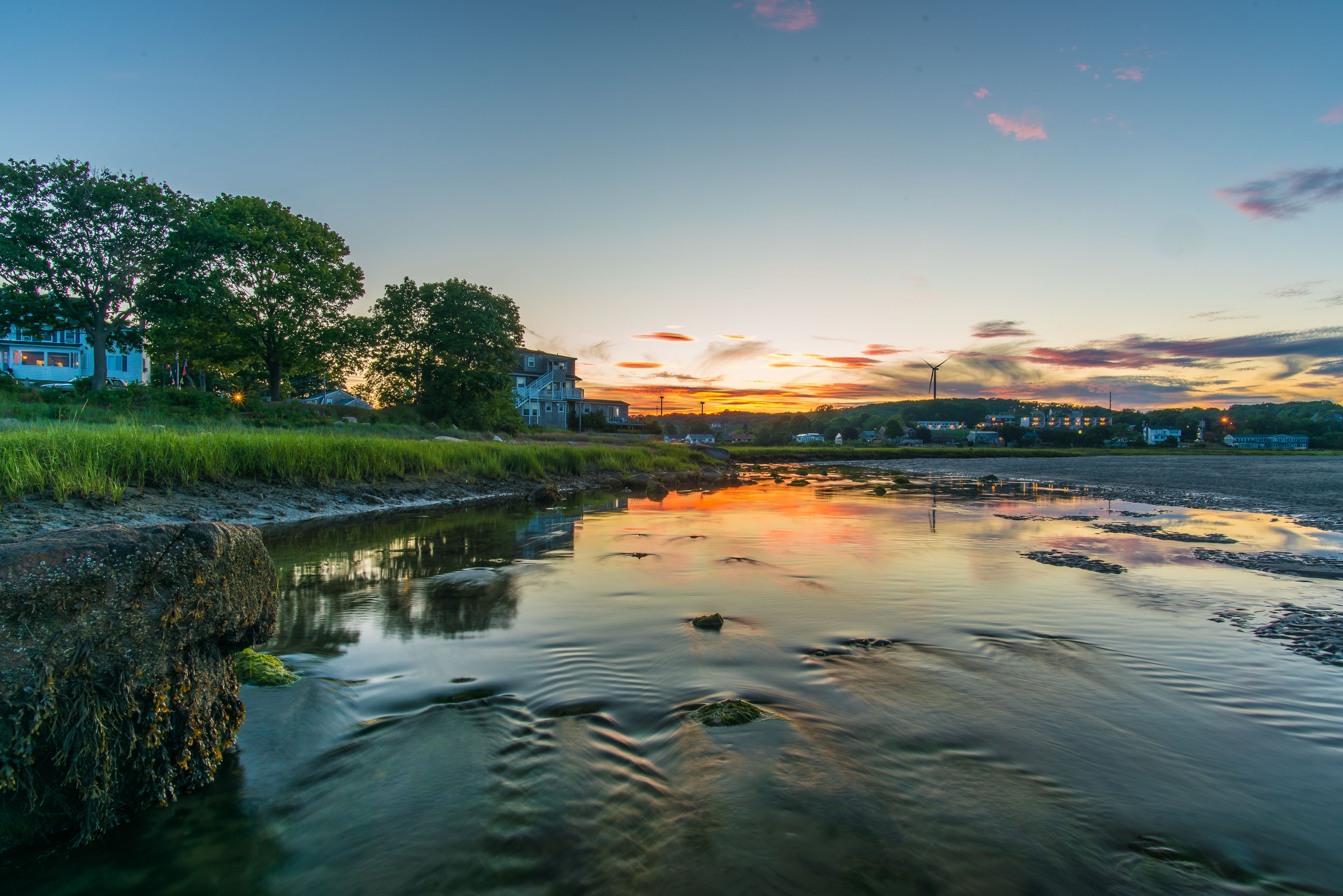 A calm river with a sunset in the background, surrounded by trees and buildings.