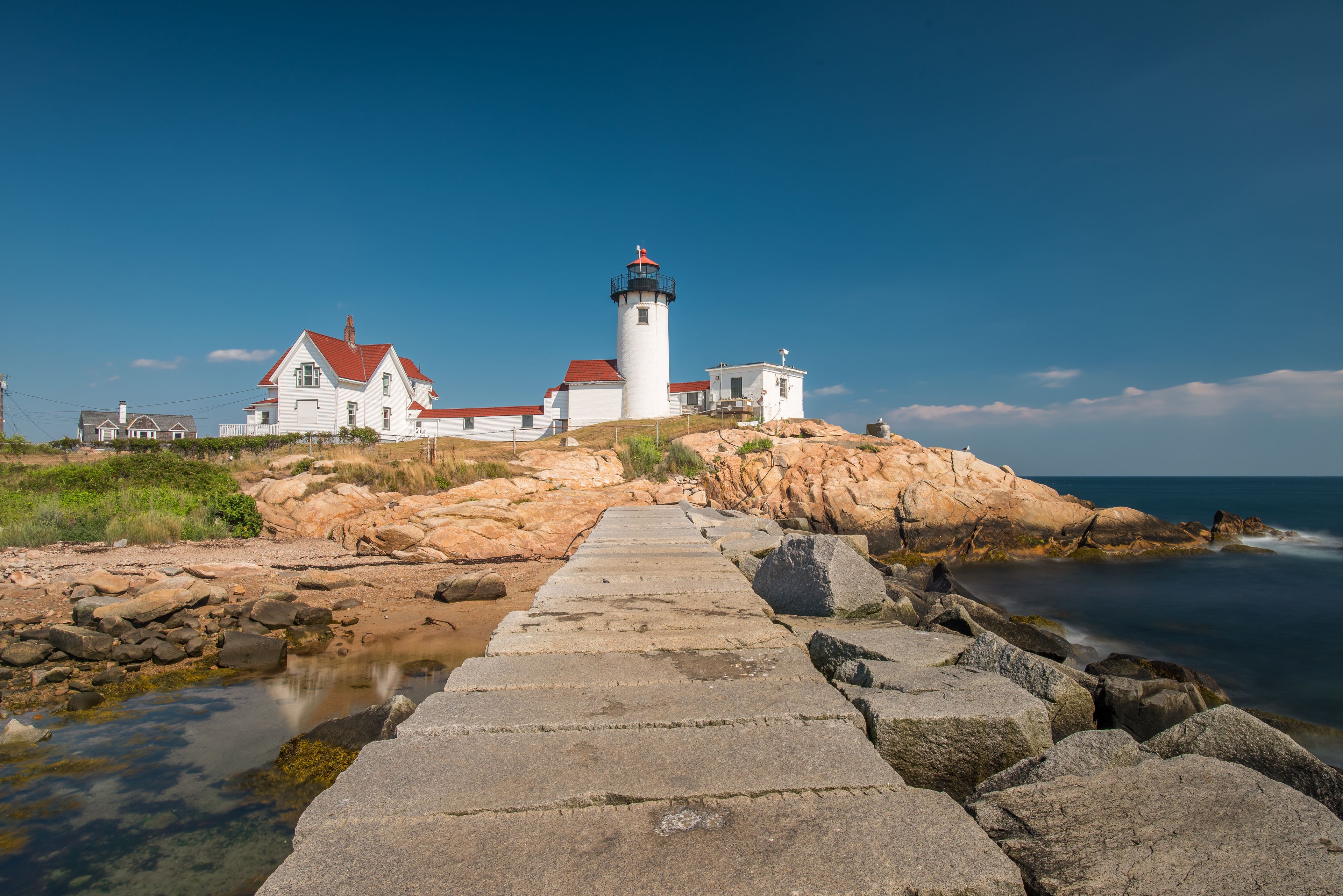 A lighthouse with a red roof, white walls, and a black top, situated on a rocky shoreline with a clear view of the ocean.