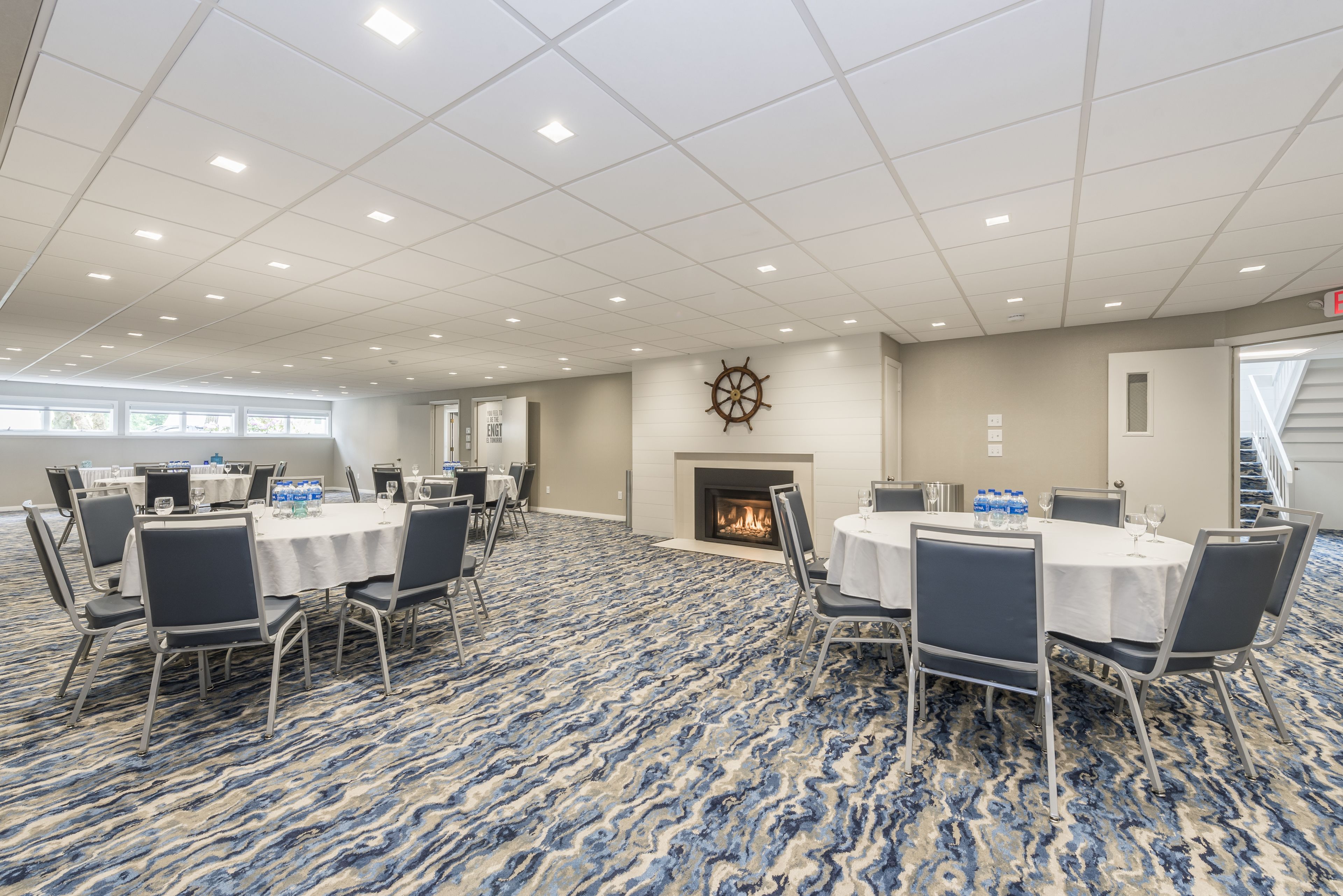 A conference room with round tables, chairs, a fireplace, and a ship's wheel decoration.