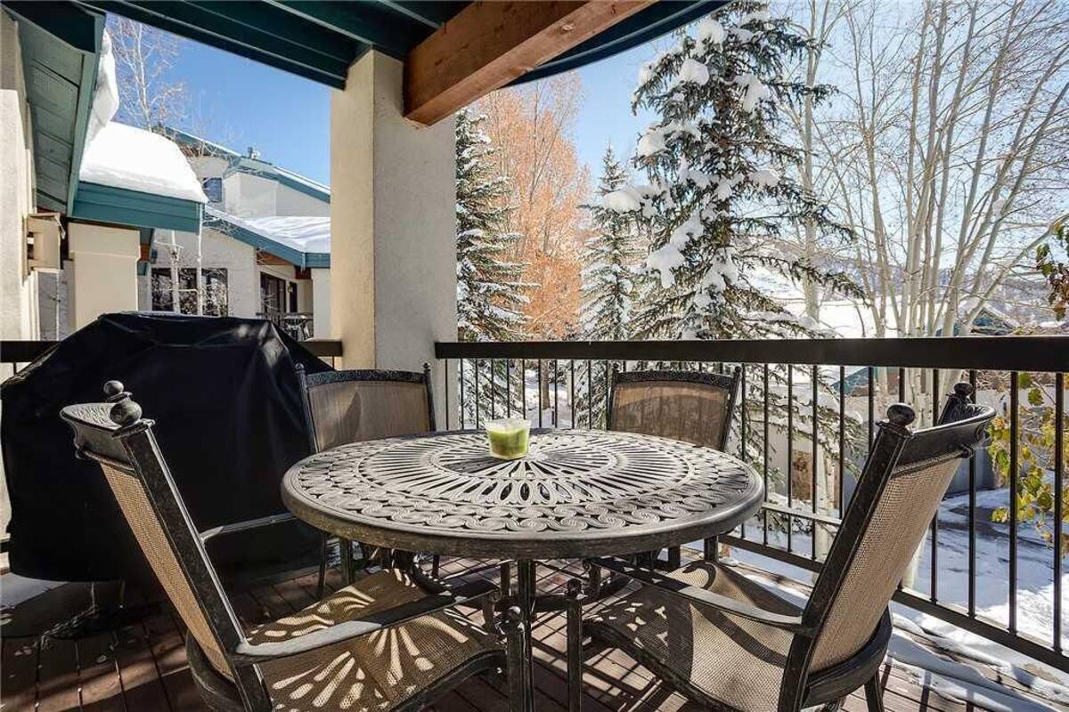 A covered outdoor patio with a round metal table and chairs, a grill, and a view of snow-covered trees.
