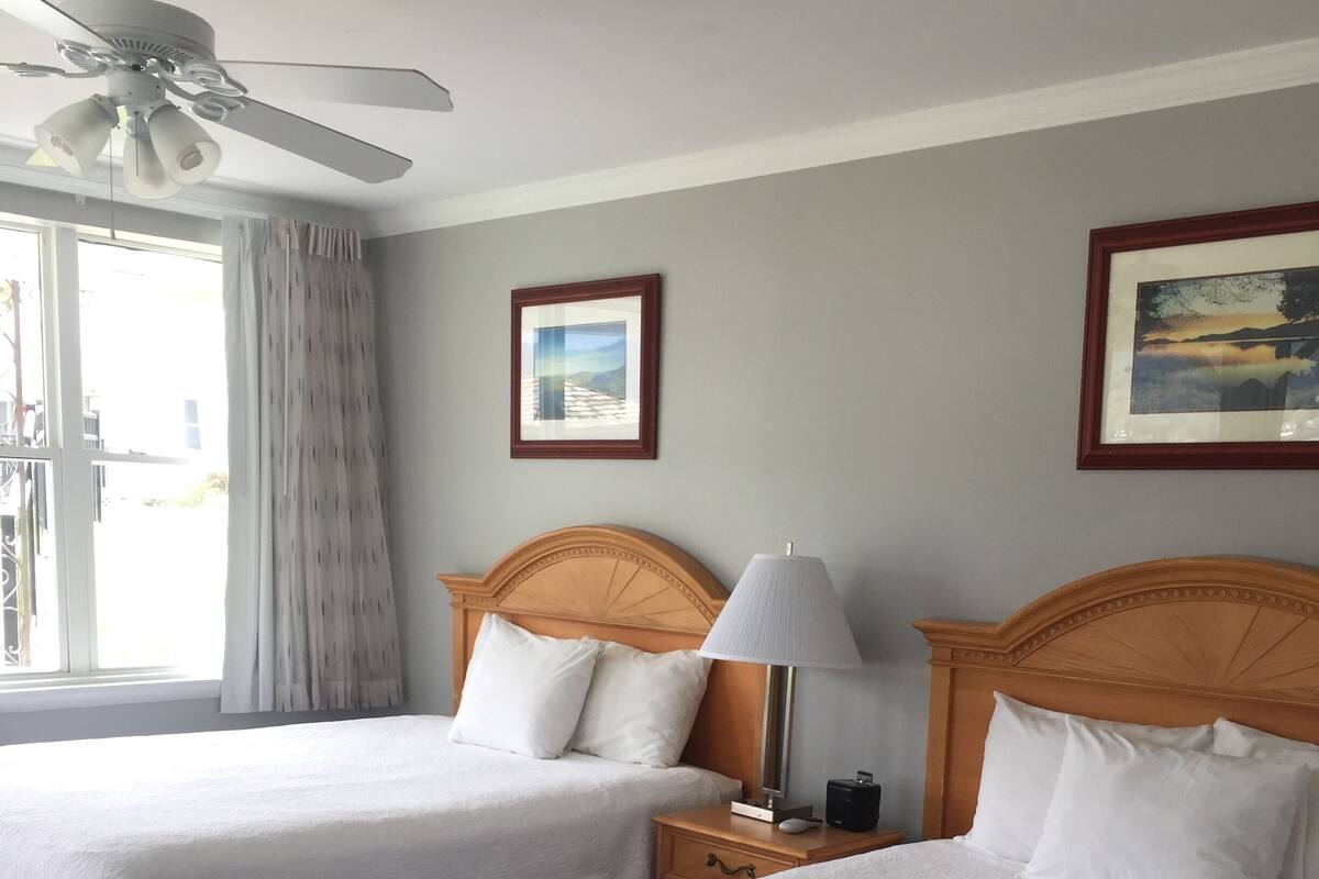 A hotel room with two beds, a ceiling fan, a nightstand with a lamp, and framed pictures on the walls.