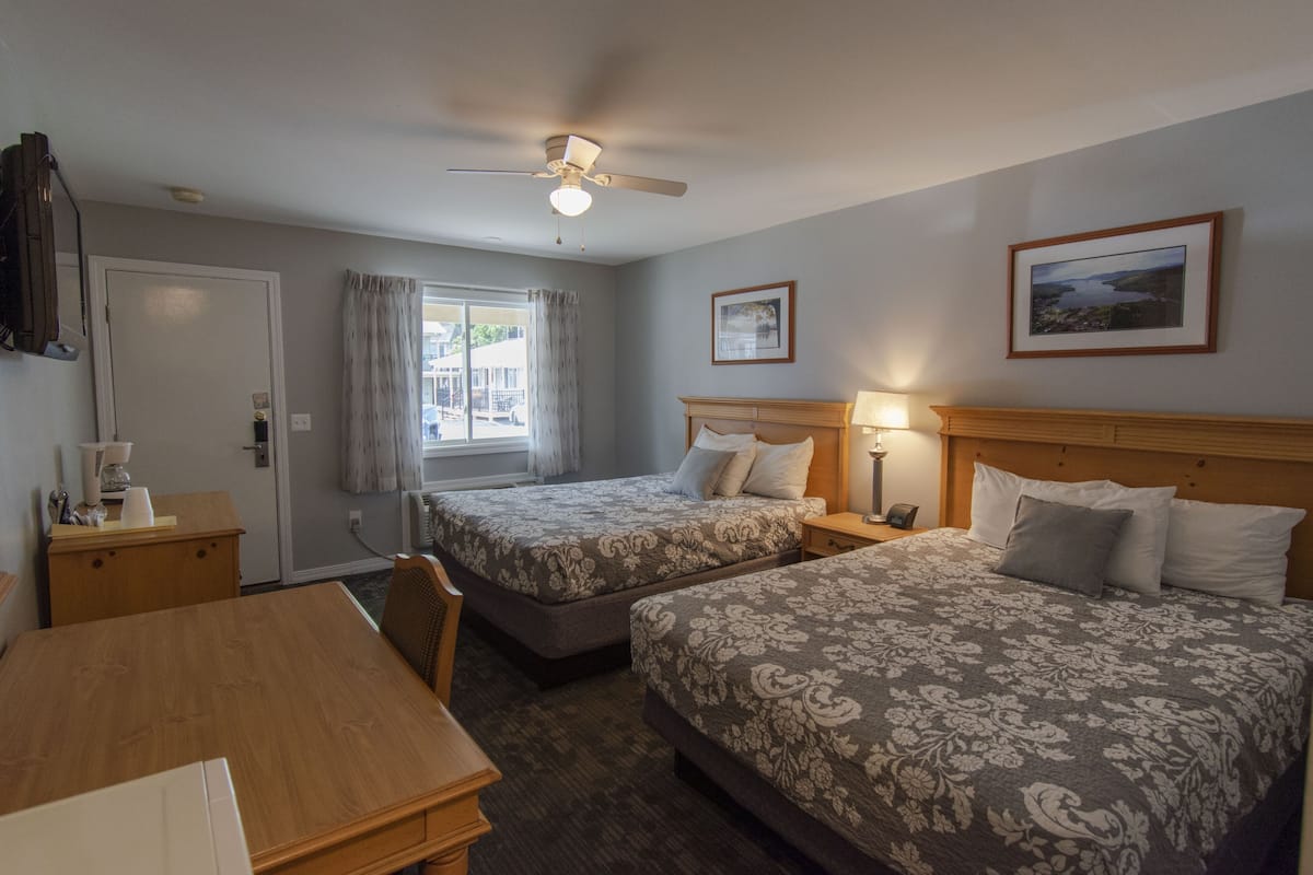 A hotel room with two beds, a wooden desk, a ceiling fan, and a window with curtains.