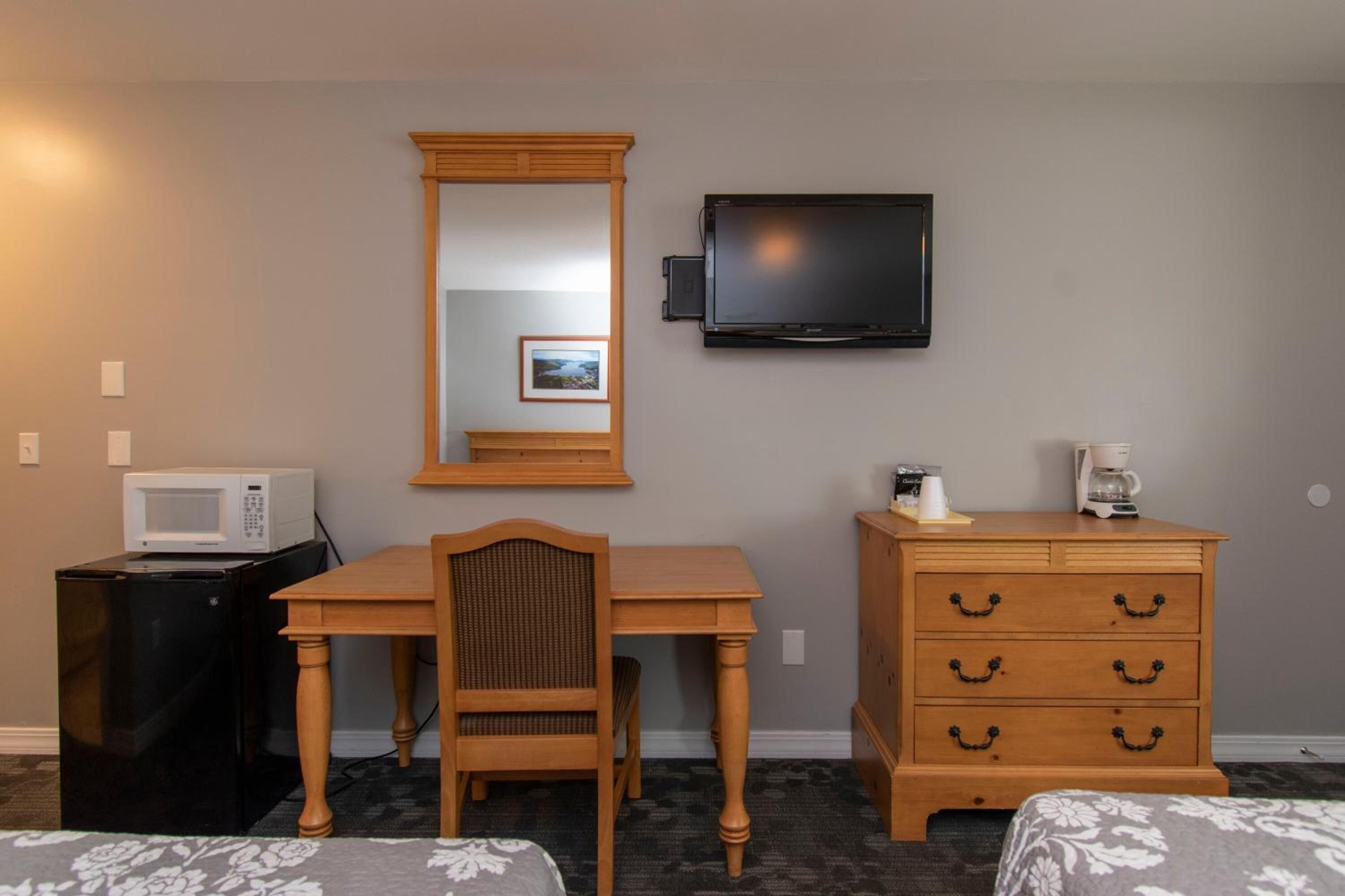 A hotel room with a wooden desk, a chair, a dresser, a television mounted on the wall, a microwave on a small table, and two beds with floral bedding.