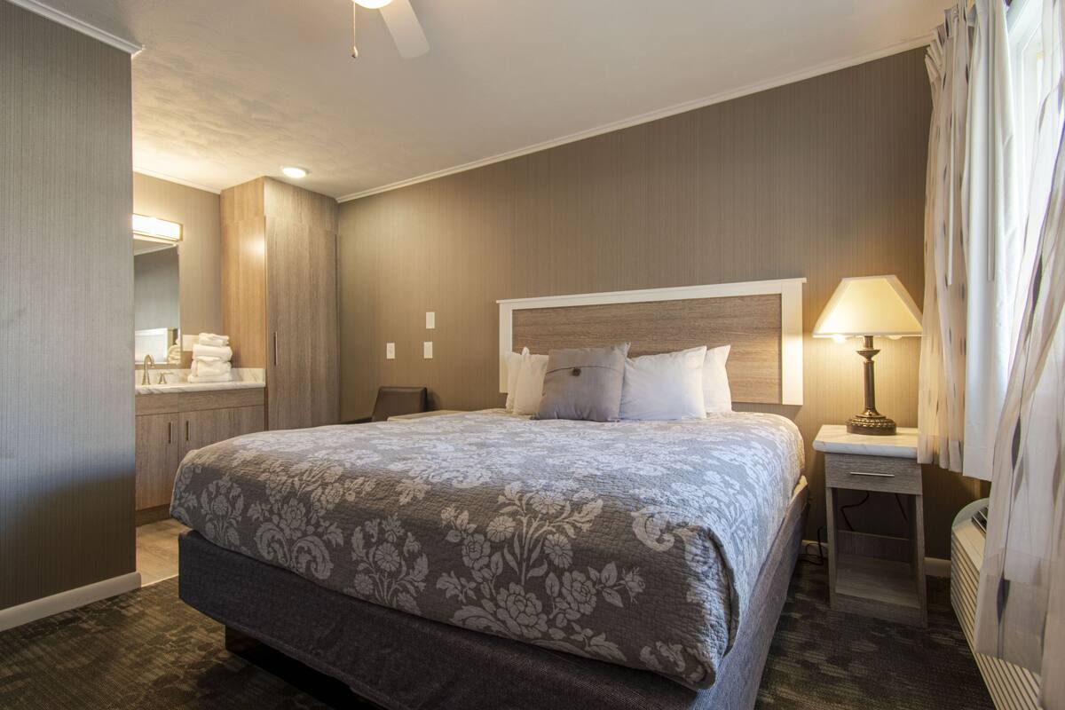 A hotel room with a large bed, a bedside table with a lamp, a nightstand, and a bathroom area with a sink and mirror.