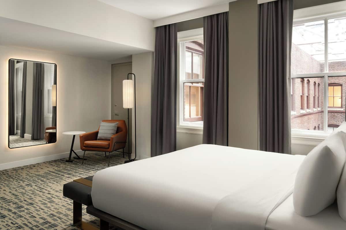 A hotel room with a large bed, a chair, a small table, a mirror, and a view of a building through the window.