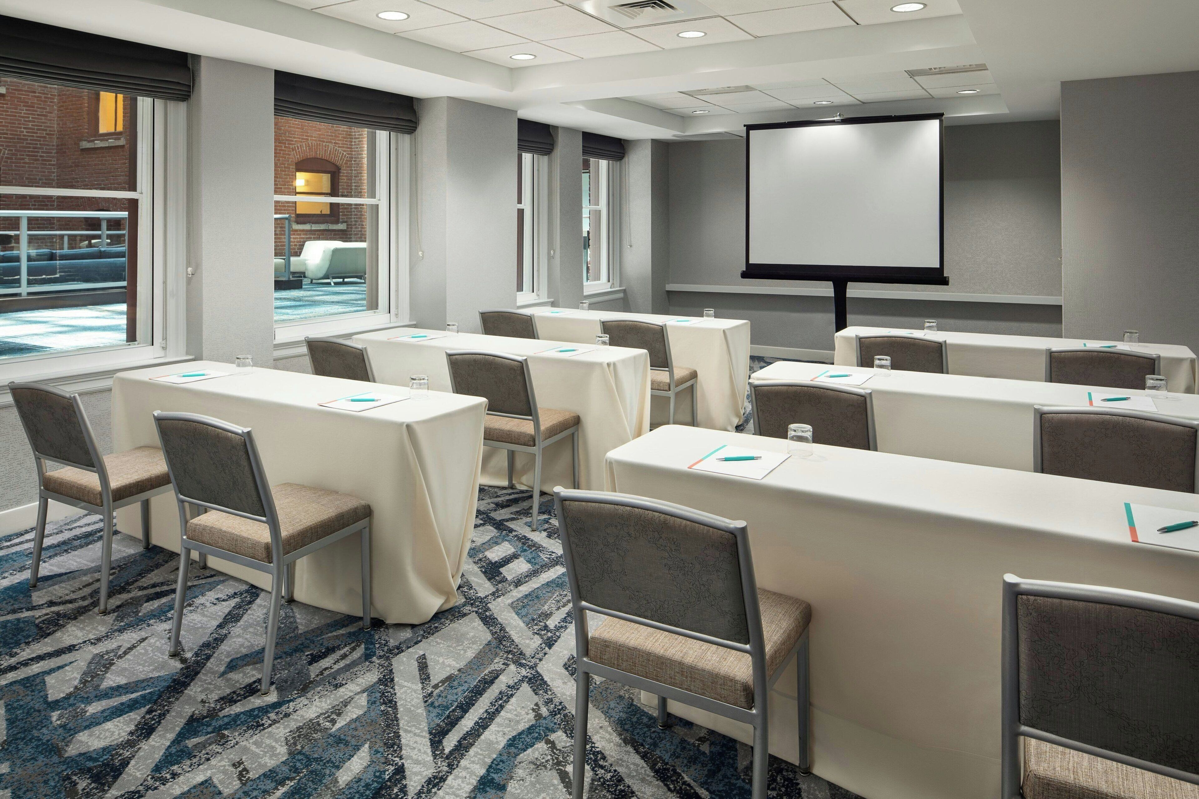 A conference room with tables, chairs, a projector screen, and large windows.