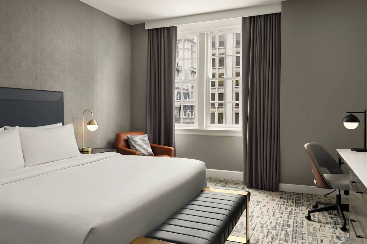 A modern hotel room with a large bed, a desk, and a view of the city through the window.