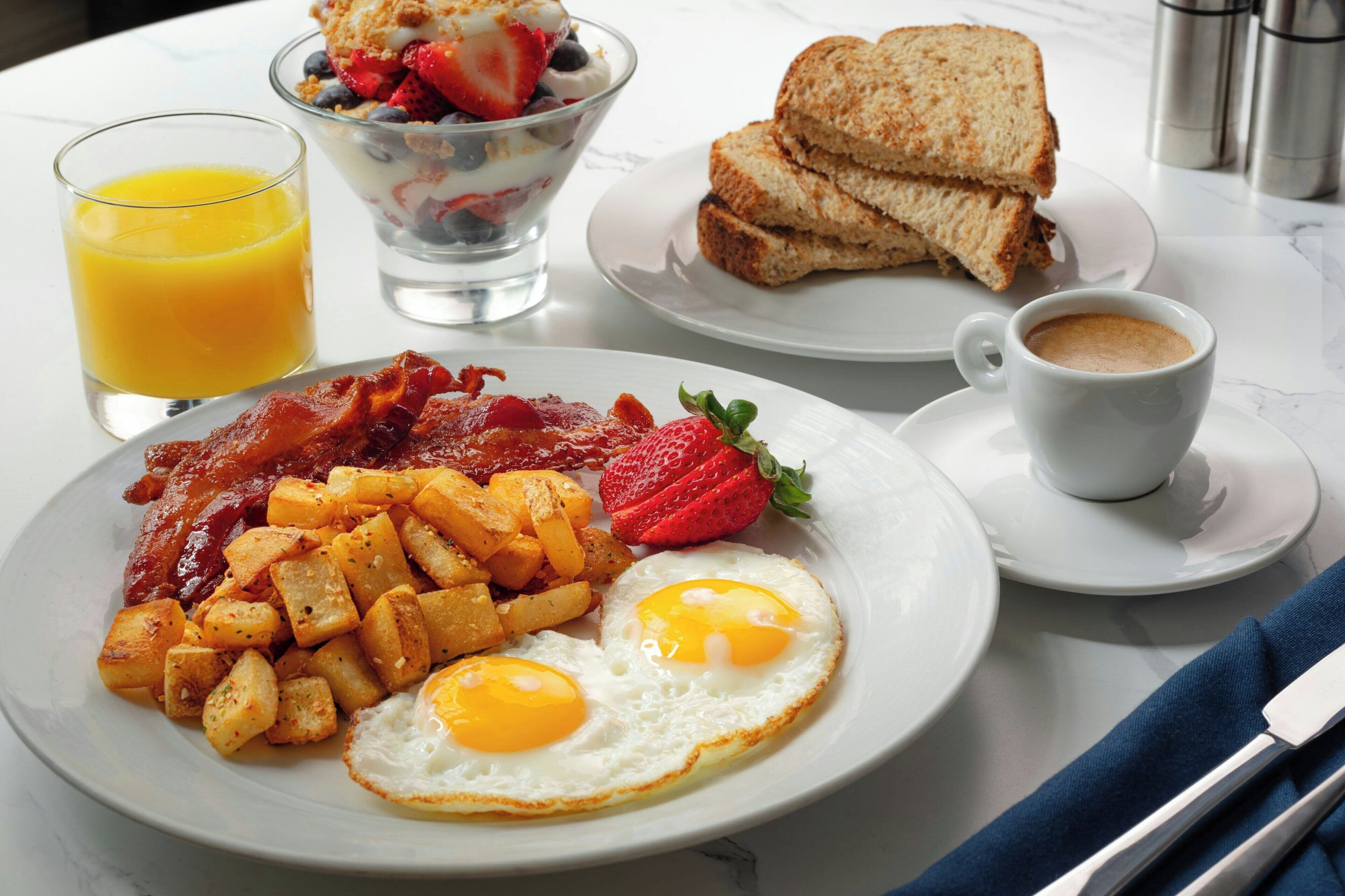 Un plato de desayuno con tocino, huevos y patatas ralladas, un vaso de jugo de naranja, un tazón de yogur con bayas, tostadas y una taza de café.