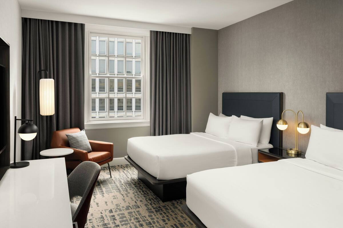A hotel room with two beds, a desk, a chair, a window with curtains, and a lamp.