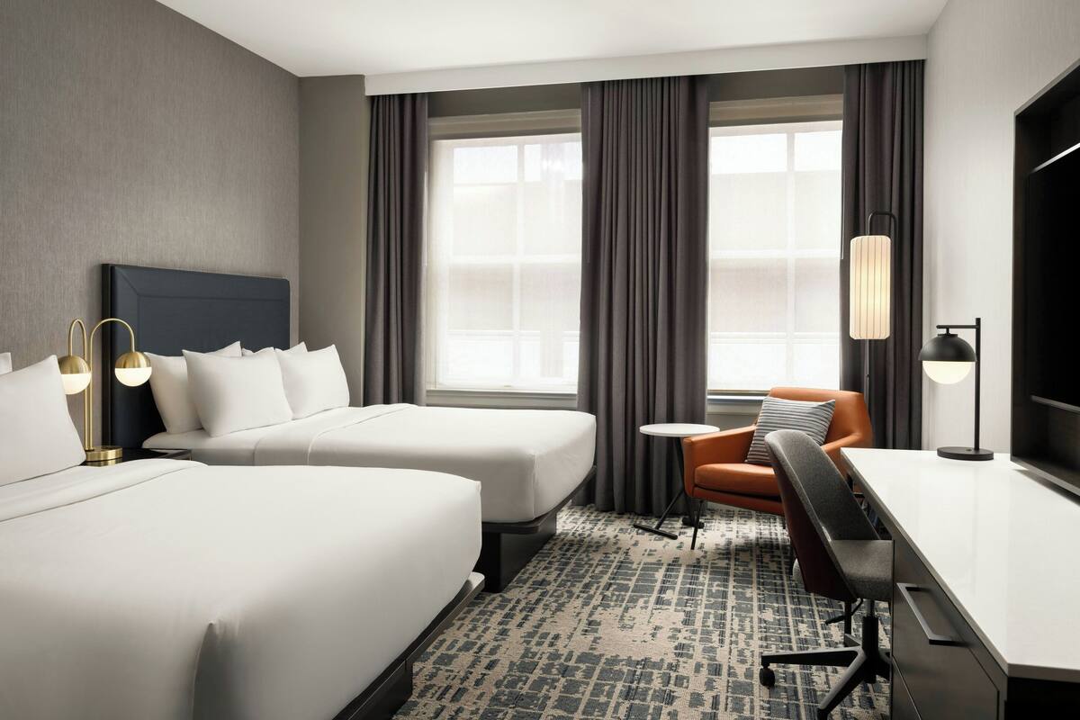 A hotel room with two beds, a desk, a chair, a TV, and a lamp.