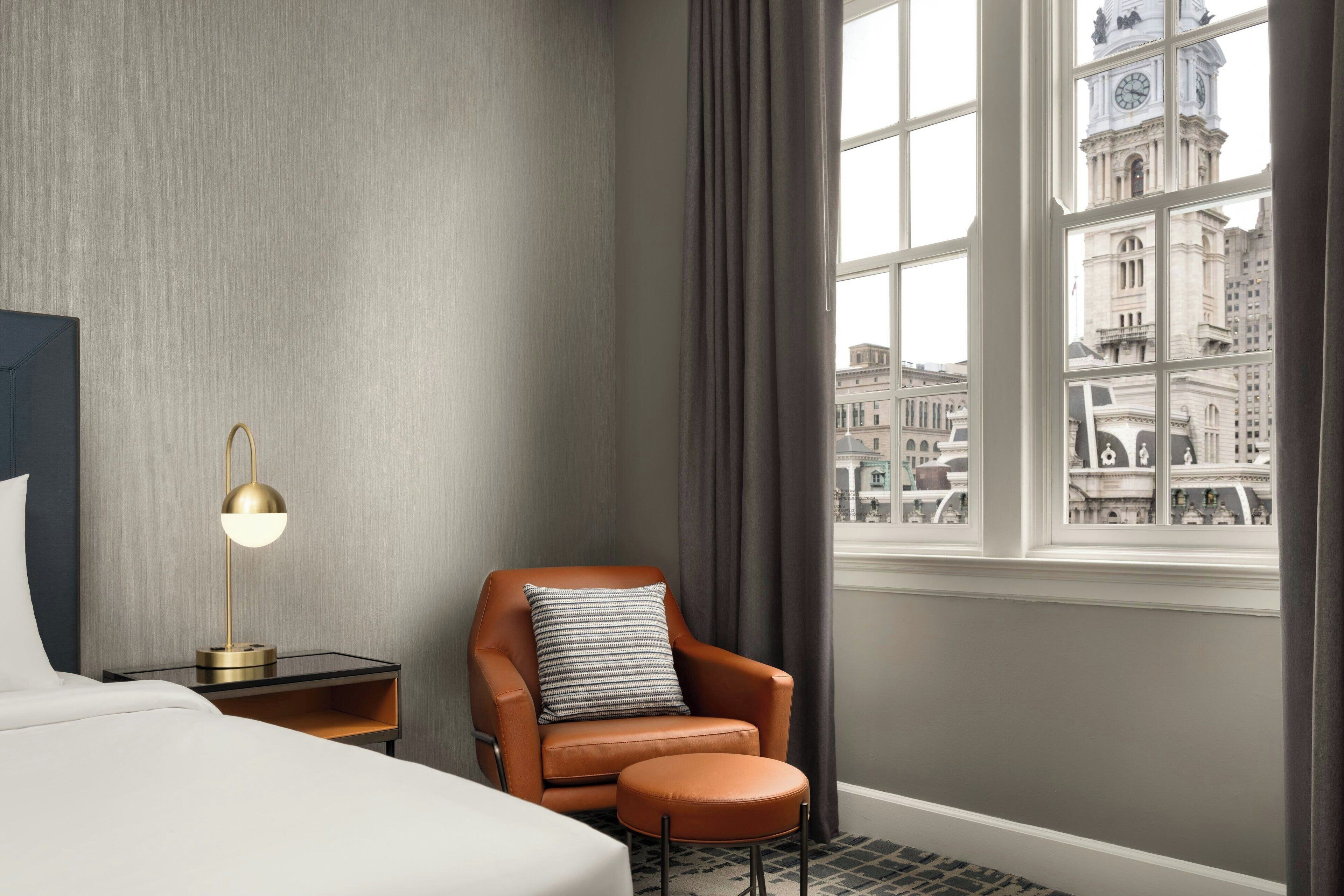 A hotel room with a bed, a leather armchair, a small ottoman, a nightstand with a lamp, and a window with a city view.