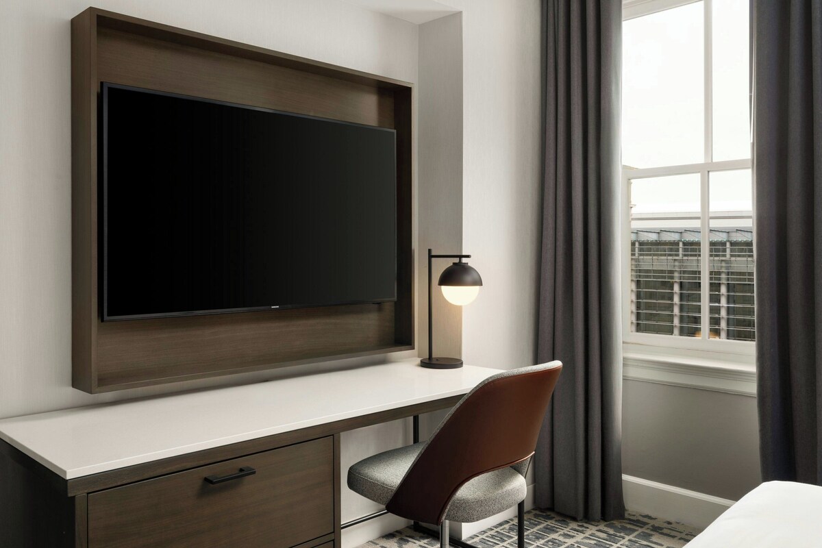 A modern hotel room with a large flat-screen TV mounted on a wooden panel above a white desk, a single chair, a floor lamp, and a window with curtains.