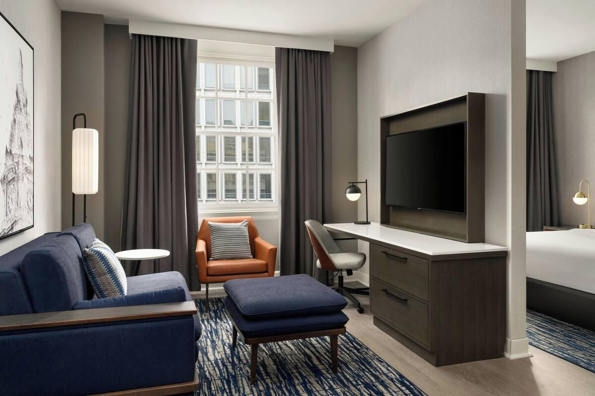 A hotel room with a sofa, armchair, ottoman, desk, TV, and a bed.