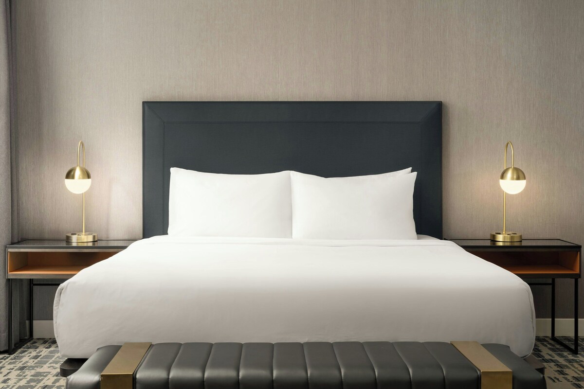 A neatly made bed with a dark headboard, two bedside tables with lamps, and a bench in front of the bed.