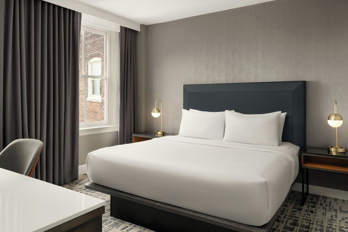A hotel room with a large bed, two bedside tables with lamps, a desk, and a window with curtains.