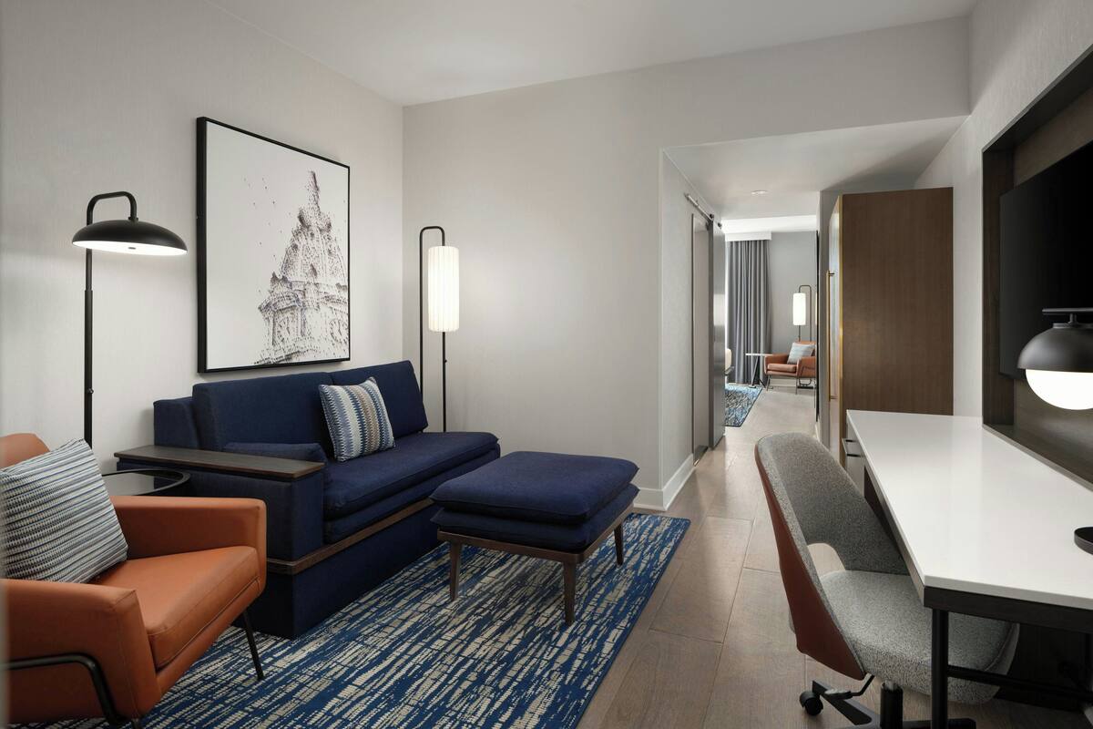 A modern hotel room with a blue sofa, orange armchair, and a desk with a chair. There is a large framed picture on the wall and a lamp on either side of the sofa.