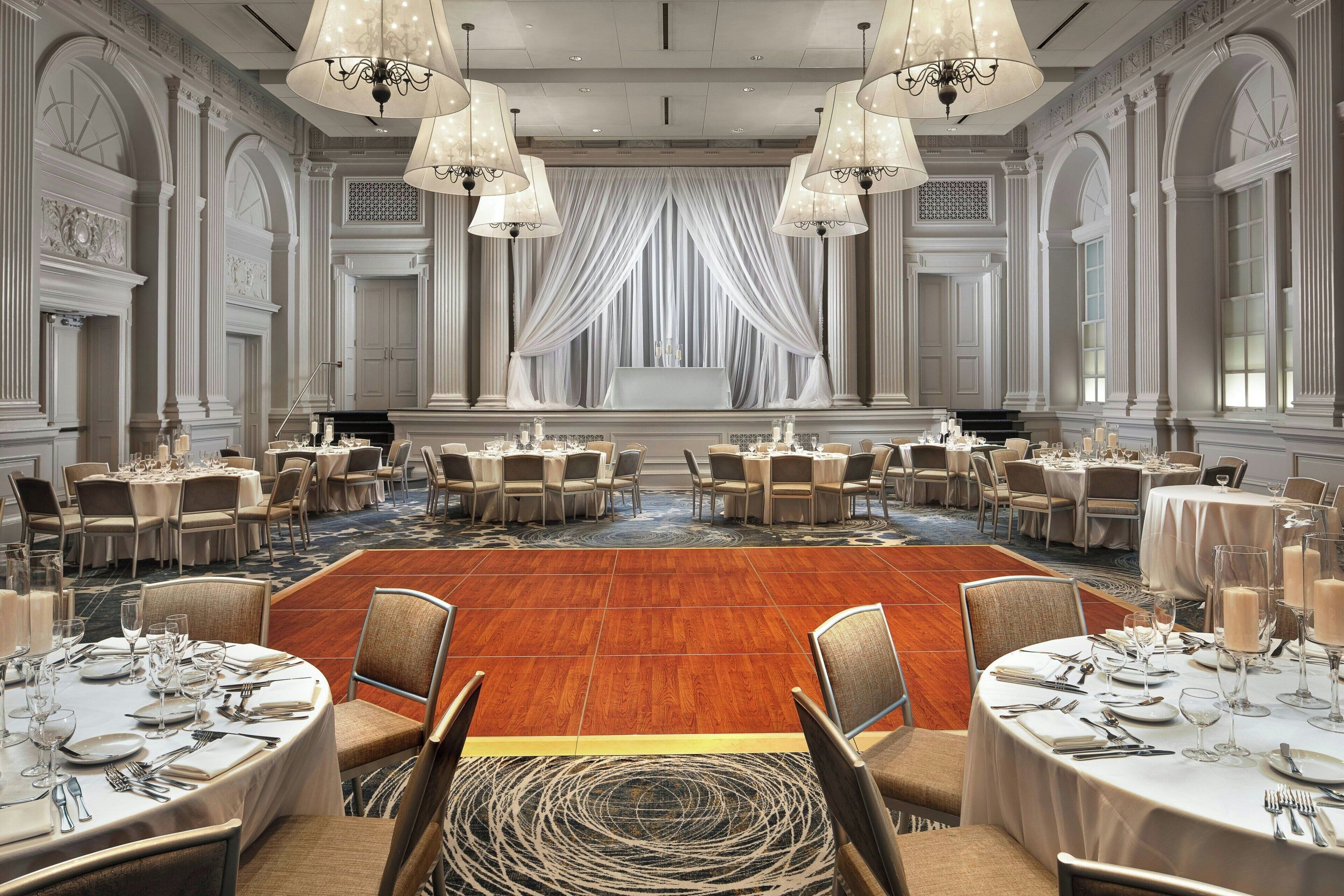 A grand banquet hall with round tables set for dining, elegant chandeliers, and a large central stage.
