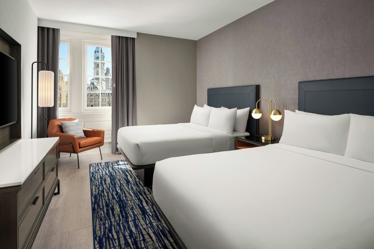 A hotel room with two beds, a desk, a chair, and a TV.