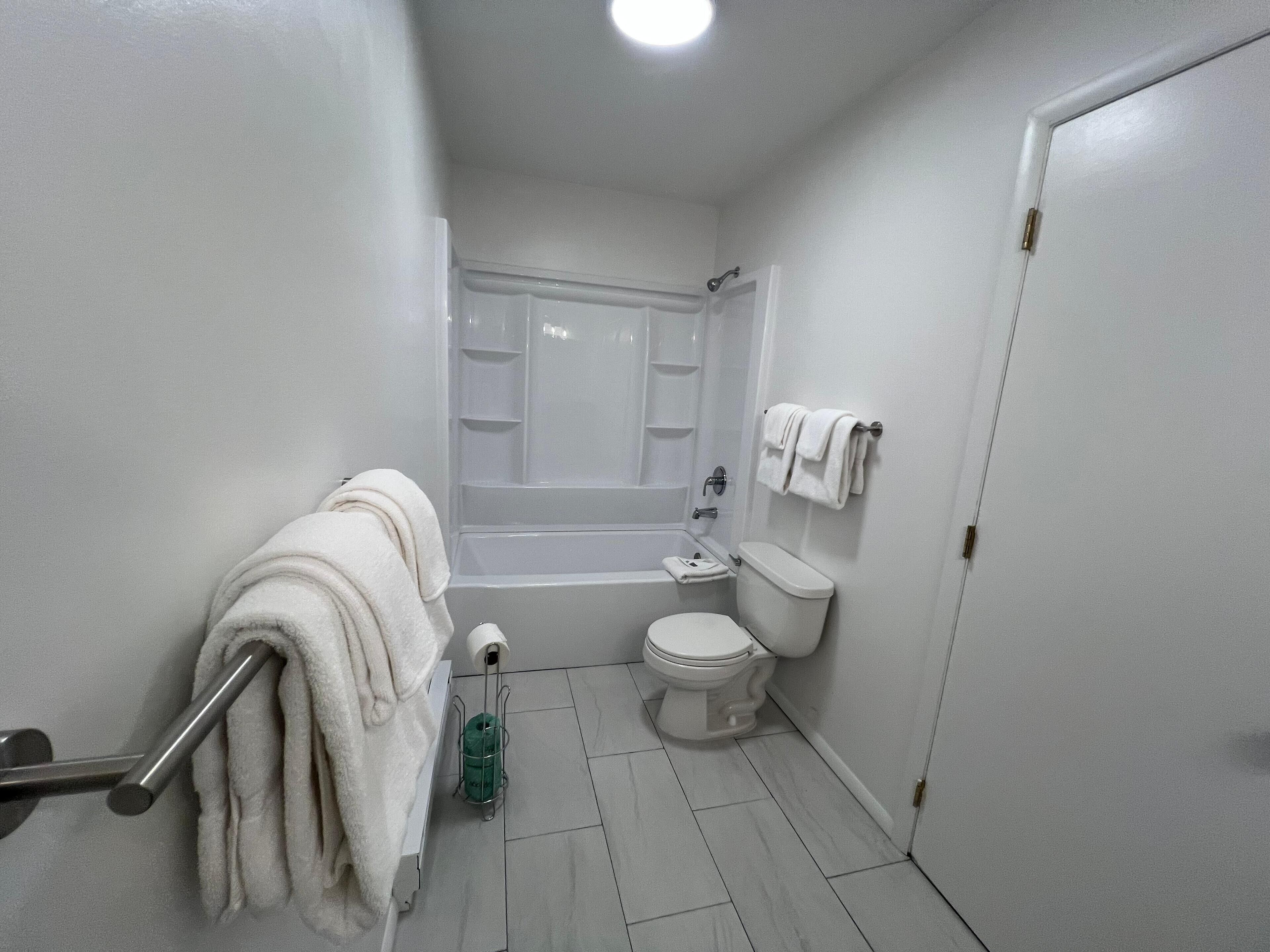 A small, clean bathroom with a toilet, sink, and bathtub. There is a towel rack with towels, a toilet paper holder, and a green cleaning spray bottle.