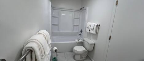 A small, clean bathroom with a toilet, sink, and bathtub. There is a towel rack with towels, a toilet paper holder, and a green cleaning spray bottle.