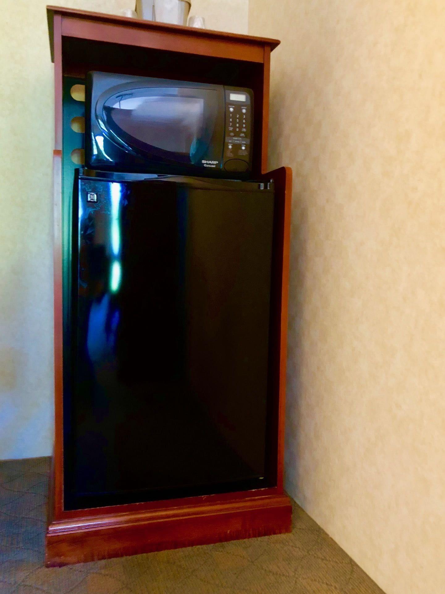 Ein schwarzer Kühlschrank mit einer Mikrowelle obenauf, platziert in einer Ecke auf einem Holzboden.